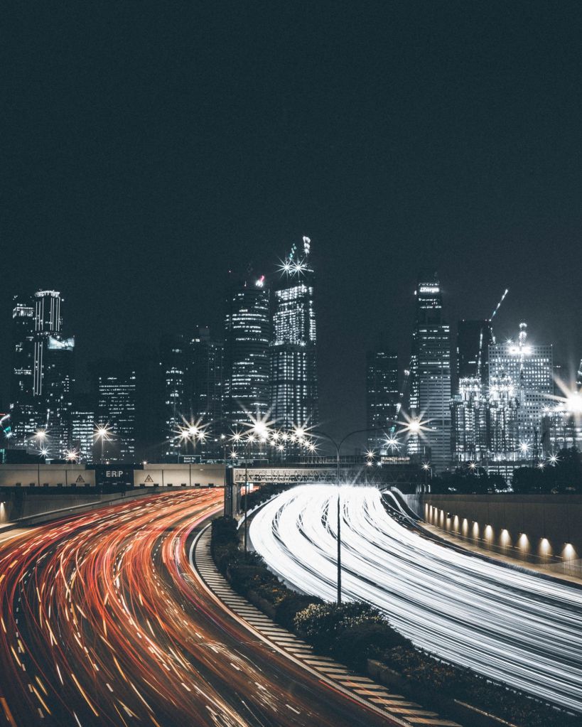 A long exposure photograph of a city at night, showcasing light trails from cars on a highway, with illuminated skyscrapers in the background.