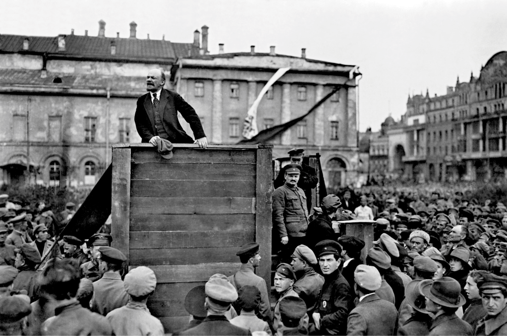 A historical black and white image of Vladimir Lenin speaking from a wooden platform in front of a large crowd during a revolutionary event.