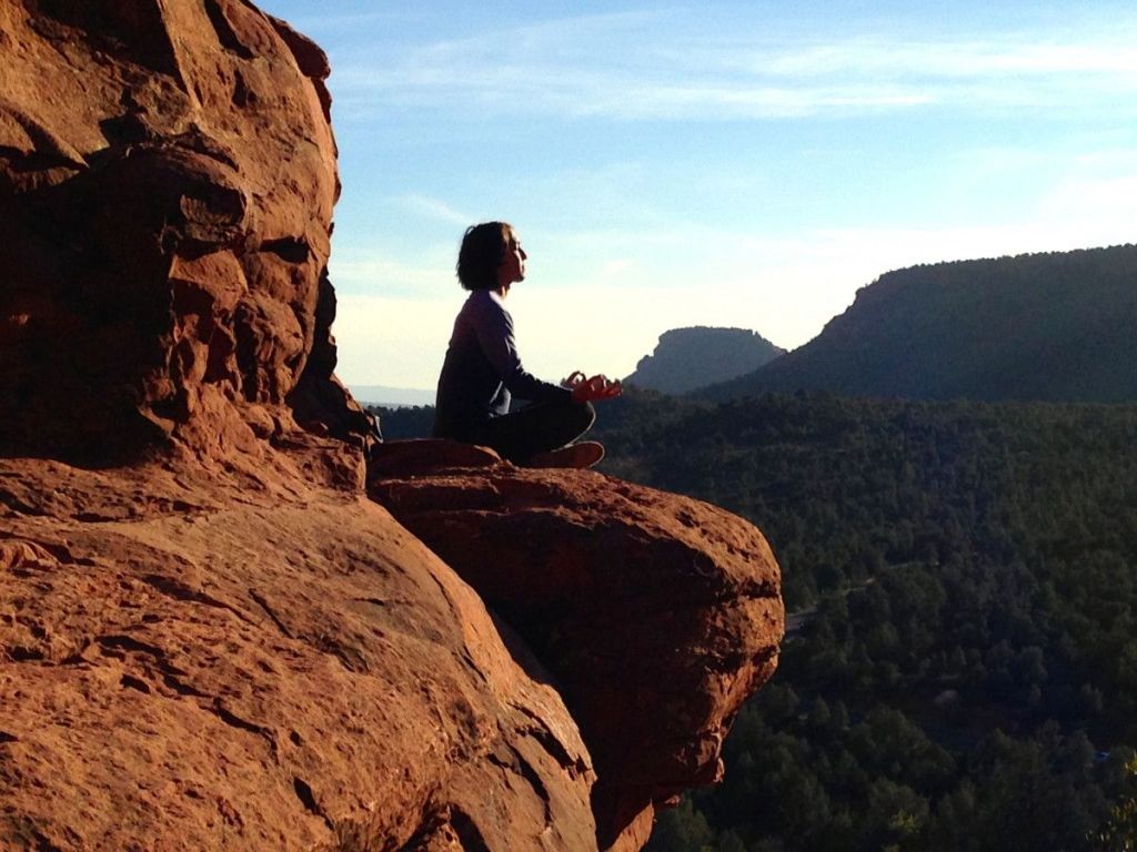 A person meditating on a rocky ledge overlooking a scenic landscape of hills and trees under a blue sky.