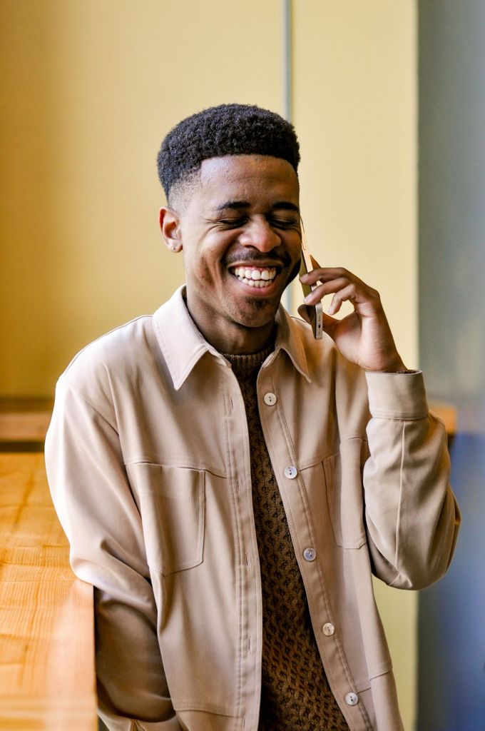 A young man laughing while talking on a smartphone, wearing a beige jacket over a brown patterned sweater, with a warm background.