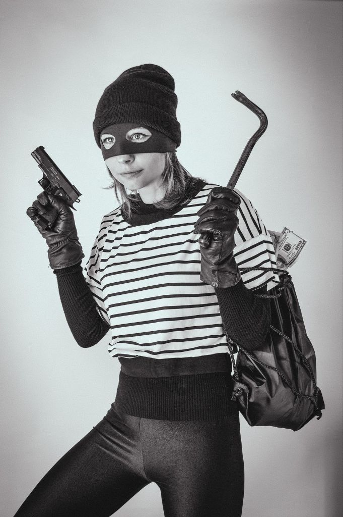 A masked woman in a striped shirt and black leggings holding a gun and a crowbar, with a bag and cash, posed to depict a thief.