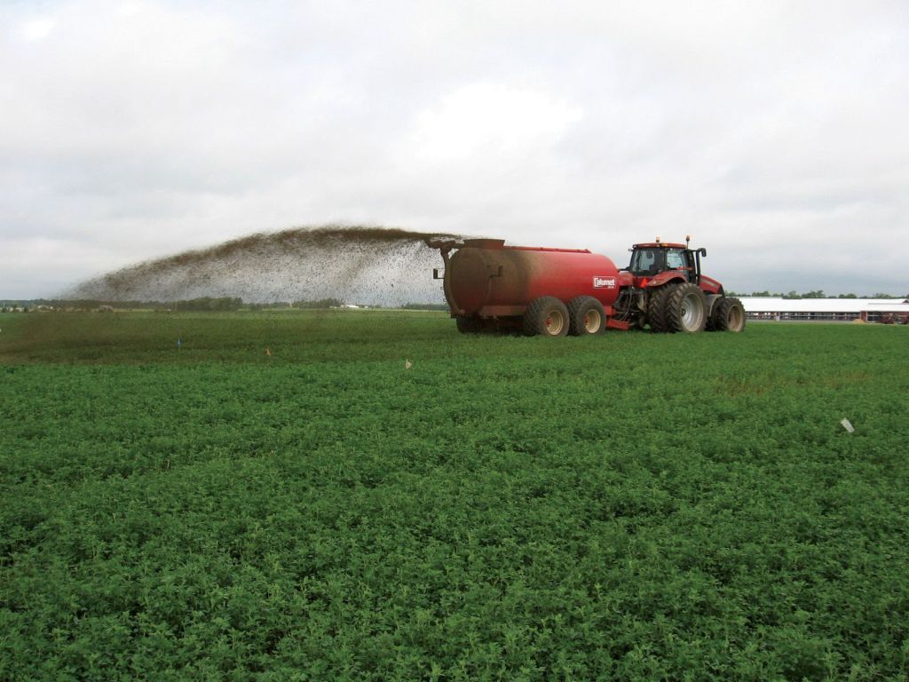 A red tractor equipped with a slurry tank spreads fertilizer across a green field under a cloudy sky.