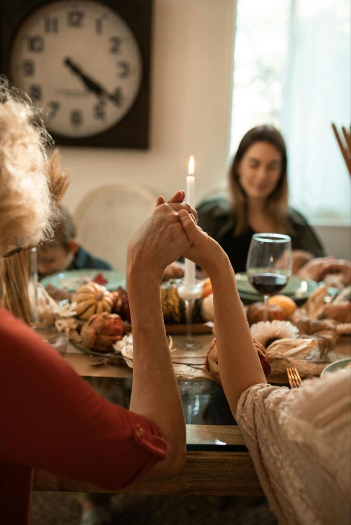 A group of people holding hands around a dinner table with a candle in the center, surrounded by seasonal decorations.