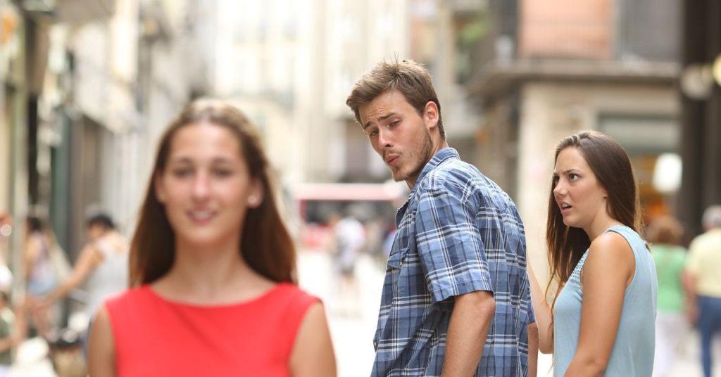 A man looks surprised while turning to glance back at a woman in a red dress, while another woman in a blue top appears displeased in a bustling street setting.